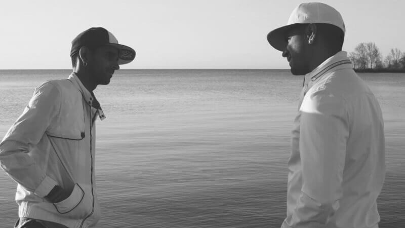 A recent photo of Vivek and Shamik standing face-to-face on the shore of a lake, smiling, both wearing baseball caps and white windbreakers.