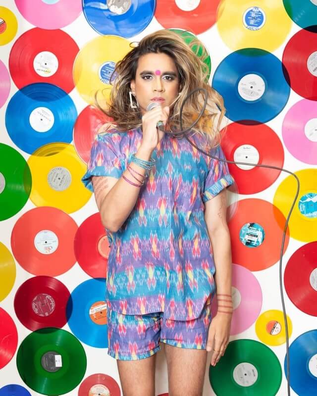 A brown-skinned trans woman with blonde-streaked hair and a red bindi, holding a microphone and standing in front of a wall covered in colourful vinyl records.