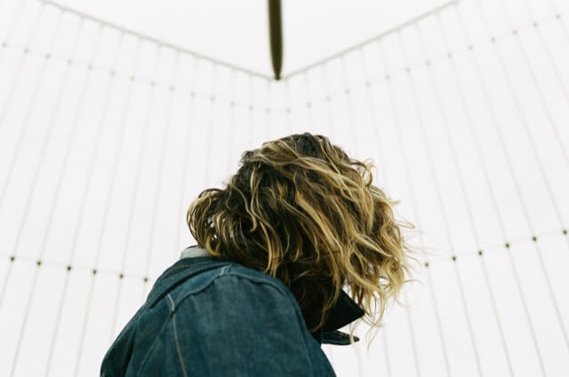A head-and-shoulders photo of a woman in a denim jacket standing on a bridge in front of an anti-suicide fence. Her face is turned away and obscured by her upturned collar and blonde-streaked black hair.
