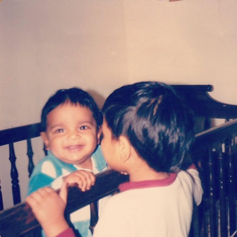 Baby Shamik standing up in his crib, grinning at the camera, while Vivek peers intently at him from a few inches away.