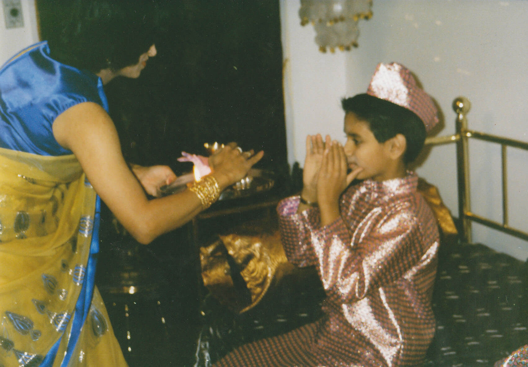 A young Vivek sits in a festive pink outfit, hands raised to her face, while her mother, in a blue and yellow sari, leans towards her with a lit lamp.