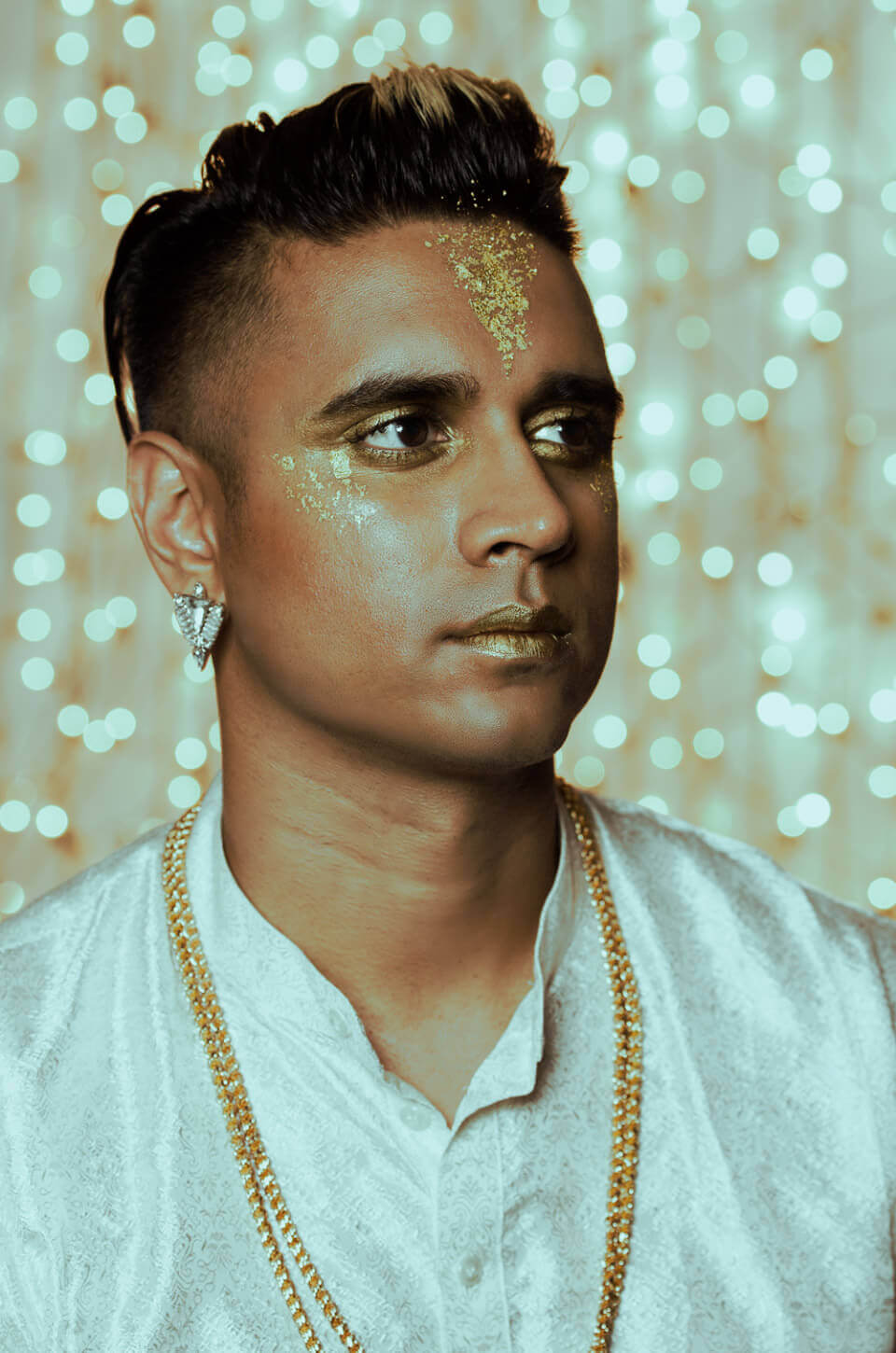 A recent head-and-shoulders photo of Vivek wearing a white tunic, gold and silver jewelry and gold makeup, in front of a glittering golden background.