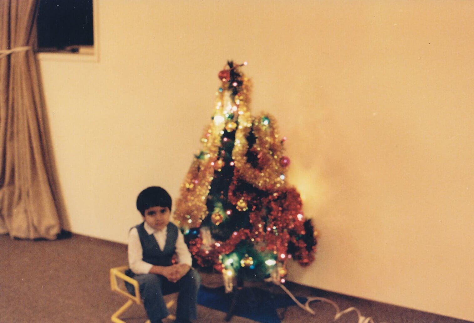 An even younger Vivek sits in front of a small, very '80s Christmas tree in a suburban living room, wearing a blue vest over a white dress shirt.