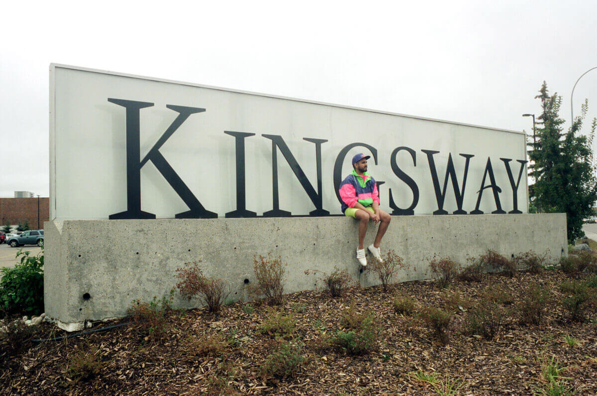 A brown-skinned person with a baseball cap and beard in a brightly-coloured windbreaker and shorts sitting on the Kingsway Mall sign in Edmonton.